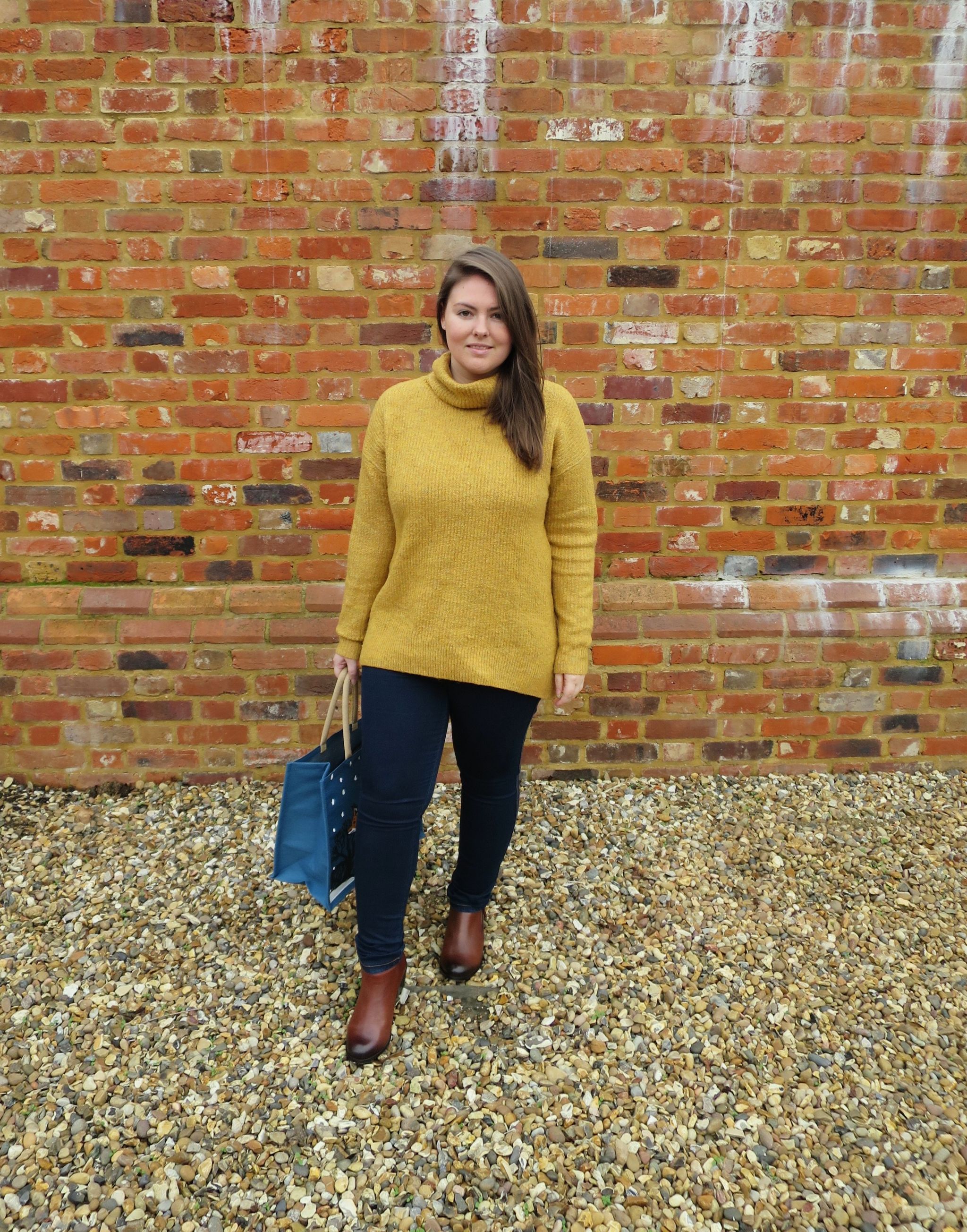 Me standing by a brick wall wearing a mustard jumper, jeans and my Vionic Shoes